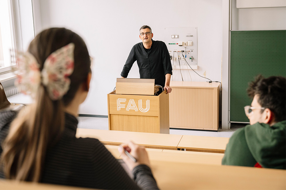 Ein Professor steht an seinem Pult im Hörsaal und redet mit seinen Studenten.