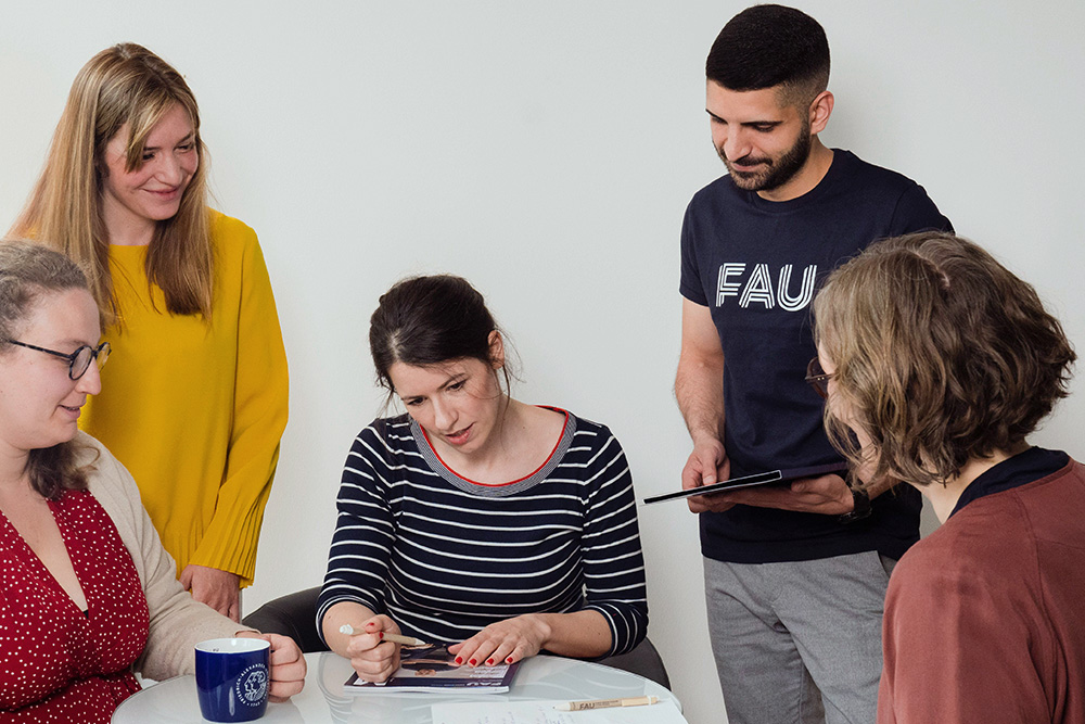 Drei Kolleginnen und Kollegen stehen um einen Tisch herum, an dem zwei weiter Kolleginnen sitzen und gerade ein Magazin besprechen.