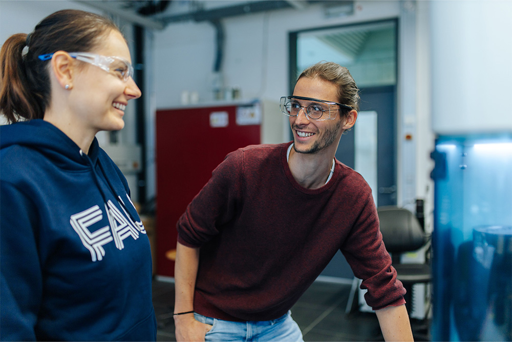 Zwei junge Forschende mit Schutzbrillen sehen sich an und lachen.