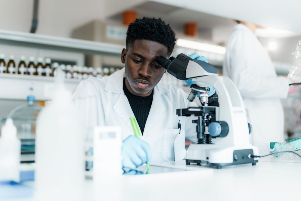 Ein Chemielaborant mit Kittel und Schutzbrille arbeitet gerade an einer Laborprobe, während ihm ein weiterer Chemielaborant mit Kittel dabei zusieht.