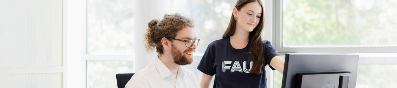 Zwei Auszubildende sitzen beziehungsweise stehen in einem hellen Büroraum der FAU vor einem PC. Die eine Person trägt ein FAU-TShirt, während die andere Person ein weißes Hemd trägt.