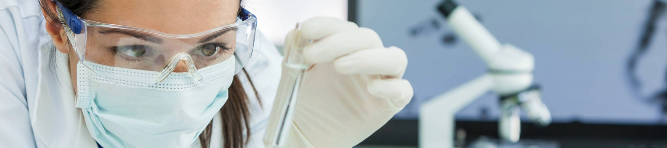 Eine Wissenschaftlerin im Labor mit Schutzbrille und Maske untersucht eine Probe. Im Hintergrund sieht man ein Mikroskop