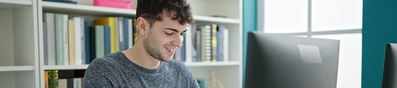 Auszubildender für Medien- und Informationsdienste sitzt vor einem Bücherregal an einem Schreibtisch und tippt etwas auf einer Tastatur