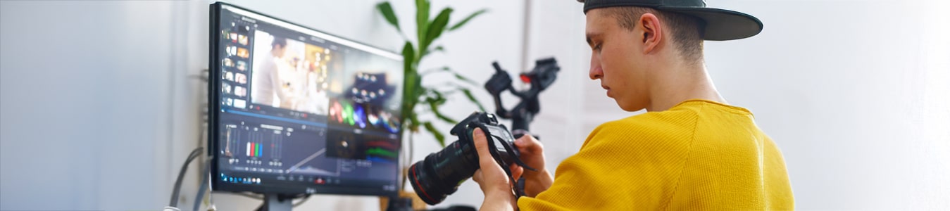 Auszubildender sitzt vor einem PC, auf dem ein Schnittprogramm geöffnet ist und betrachtet währenddessen Bilder auf einer Kamera, die er in der Hand hält