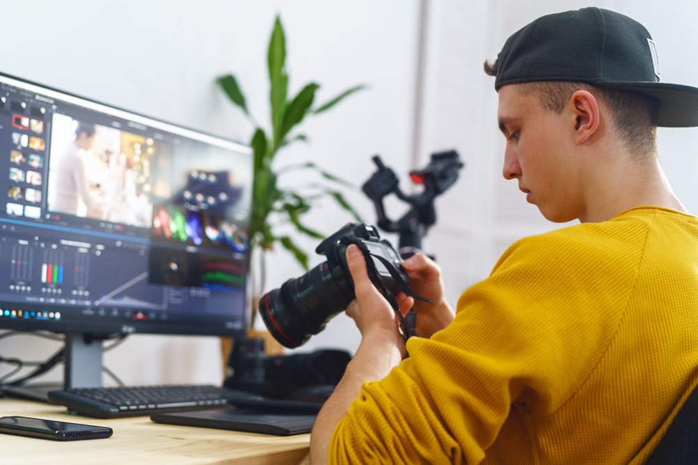 Ein Mediengestalter sitzt vor einem PC, auf dem ein Schnittbearbeitungsprogramm geöffnet ist, während er Bilder auf einer Kamera betrachtet.