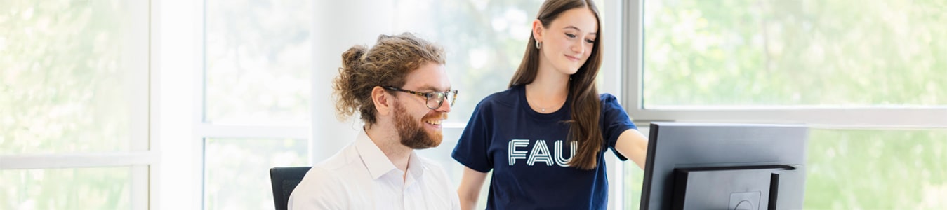 Zwei Auszubildende sitzen beziehungsweise stehen in einem hellen Büroraum der FAU vor einem PC. Die eine Person trägt ein FAU-TShirt, während die andere Person ein weißes Hemd trägt.