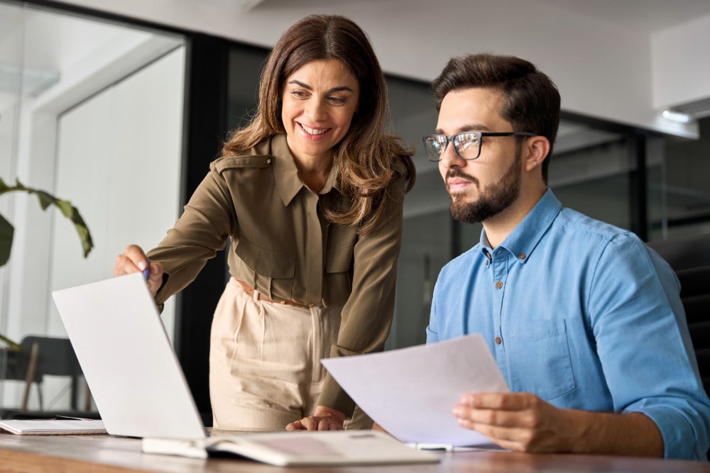 Eine weiblich und eine männlich gelesene Person besprechen Unterlagen im modernen Büro am Laptop