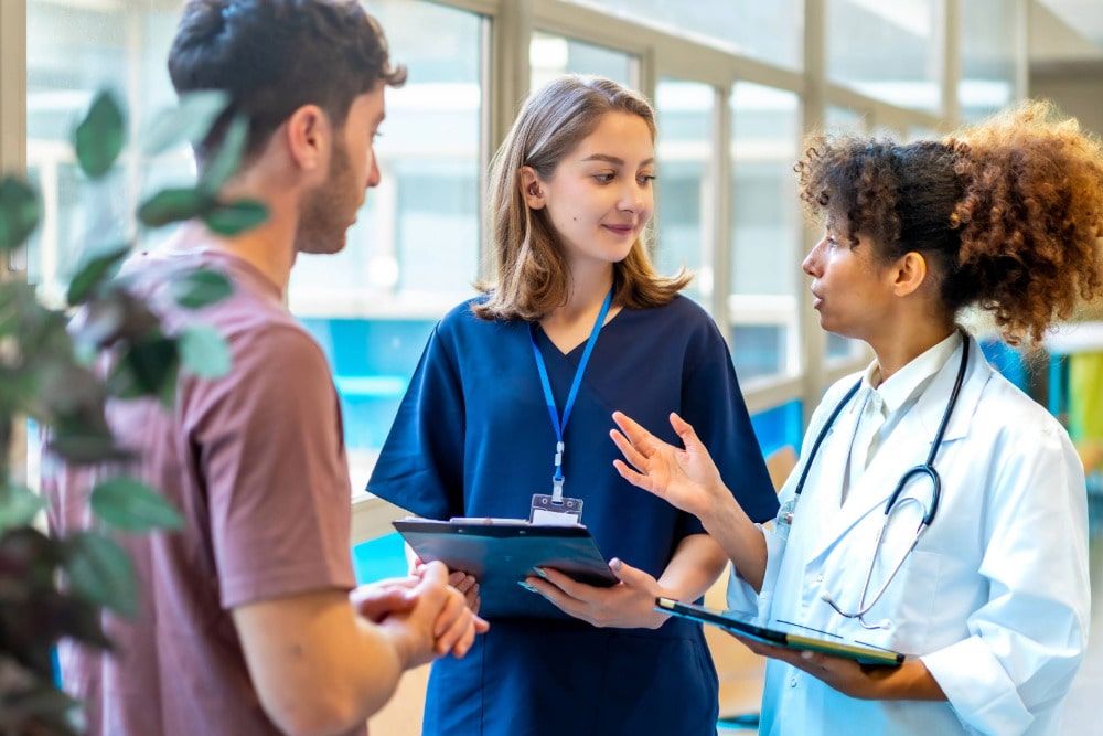 Eine Medizinische Fachangestellte befindet sich im Gespräch mit einem Patienten und einer Ärztin.