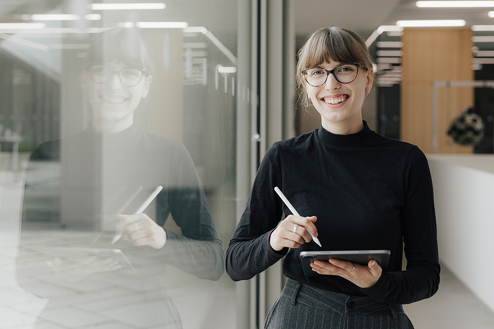 Weiblich gelesene Person steht an einem großen Glasfenster in in einem modernen Bürogebäude und lächelt in die Kamera, während sie Tablet und Stift in der Hand hält.