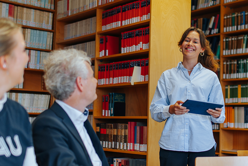 Drei Personen befinden sich in einer Bibliothek. Zwei Personen sitzen und sehen zu einer dritten, stehenden Person, die in die Kamera lächelt. Sie trägt eine gestreifte Bluse und hält ein Tablet in der Hand.