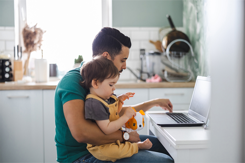 Männlich gelesene Person mit einem Kind auf dem Arm sitzt in einer Küche an ihrem Laptop.