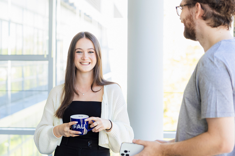 Zwei Personen stehen in einer hellen, modernen Arbeitsumgebung. Die eine Person hält eine Tasse mit dem Logo der Friedrich-Alexander-Universität Erlangen-Nürnberg in der Hand und lächelt in die Kamera. Die andere Person hält ein Smartphone.