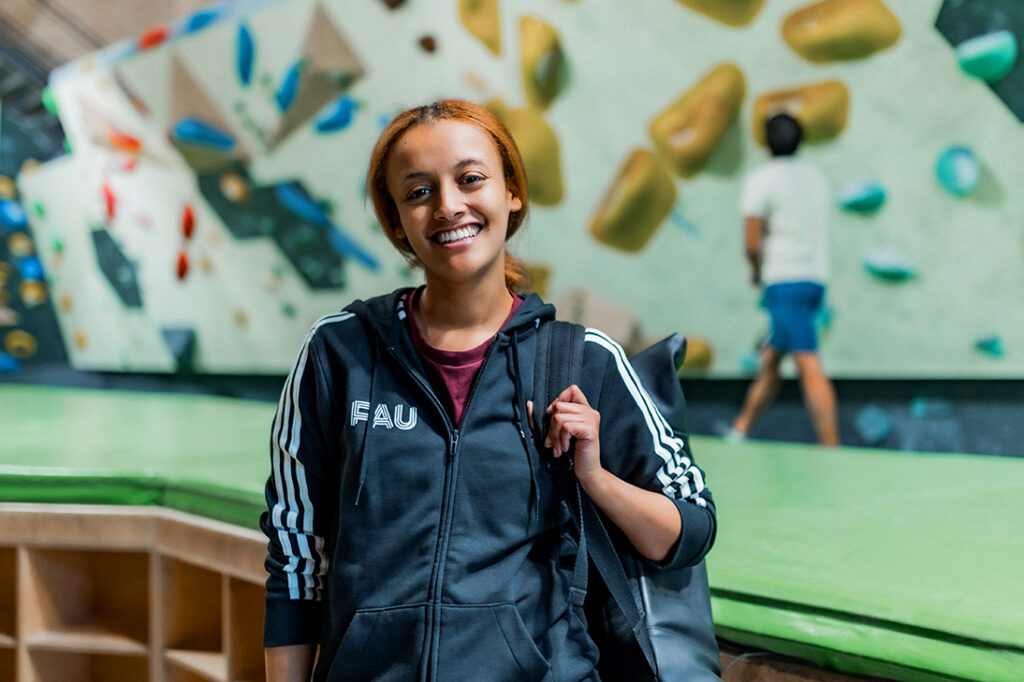 Weiblich gelesene Person steht in einer Boulderhalle und lächelt direkt in den Kamera. Sie trägt einen schwarzen Rucksack und eine schwarz Zip-Jacke mit dem Logo der Friedrich-Alexander-Universität Erlangen-Nürnberg.