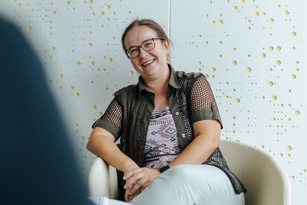 Weiblich gelesene Person sitzt in einem Sessel und lacht ihr Gegenüber an. Sie trägt Brille und ein T-shirt, dazu eine Weste.