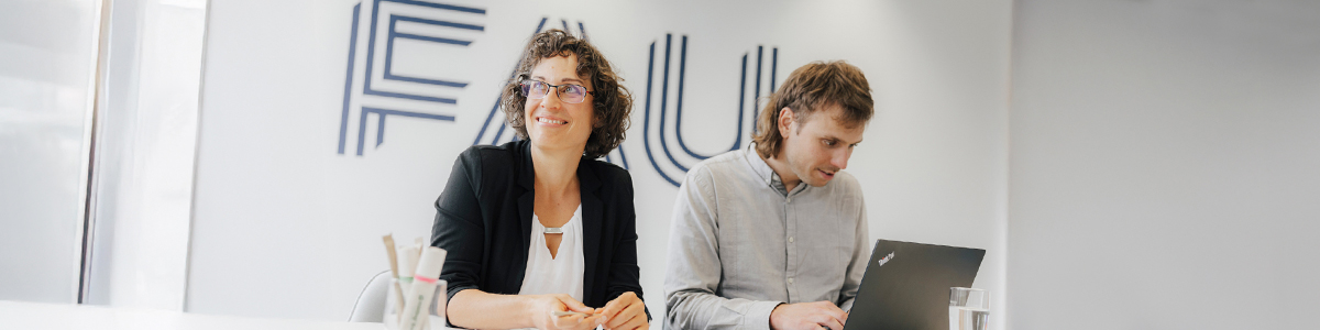 Zwei Personen sitzen an einem Schreibtisch in einem weißen Raum. Im Hintergrund ist das Logo der Friedrich-Alexander-Universität Erlangen-Nürnberg zu sehen. Beide Personen tragen Business-Kleidung. Die eine Person schreibt etwas auf ihrem Laptop, die andere Person blickt lächelnd in Richtung Fenster.