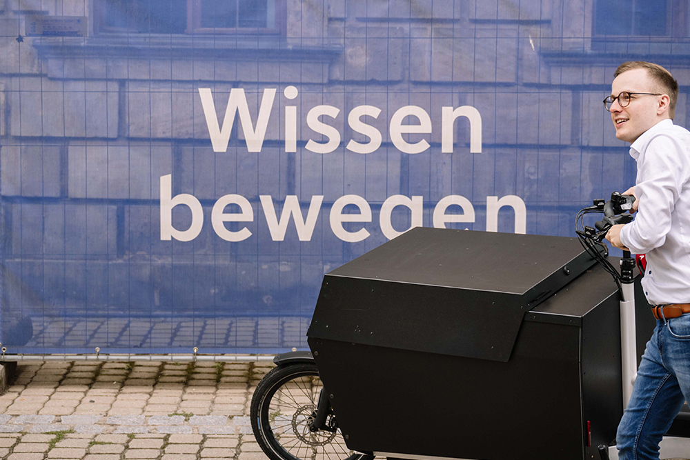 Eine männliche gelesene Person schiebt ein elektrifiziertes Lastenrad von rechts nach links ins Bild. Im Hintergrund sieht man ein Banner mit der Aufschrift "Wissen bewegen" in weißer Schrift auf blauem Untergrund.