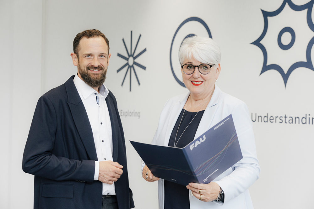 Zwei Personen stehen in einem Raum, auf dem an der Wand ein Wandtattoo mit den Worten "Exploring" und "Understanding" angebracht ist. Beide Personen sind im Business-Stil gekleidet und lächeln in die Kamera. Die rechte Person hält eine Mappe mit dem Logo der Friedrich-Alexander-Universität Erlangen-Nürnberg in der Hand.