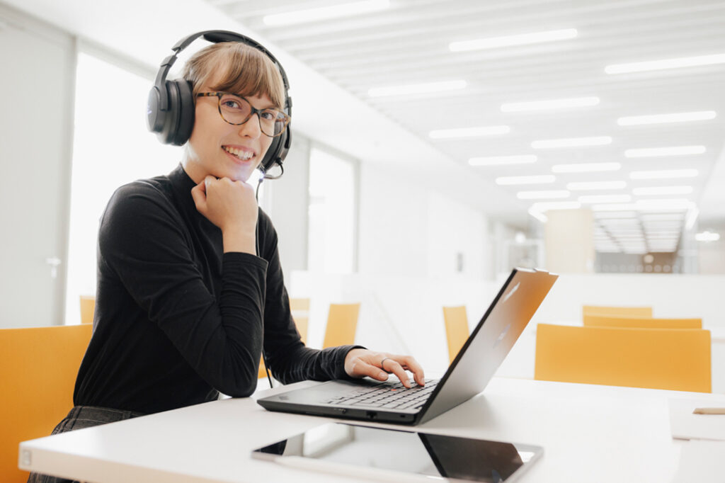 Eine weiblich gelesene Person sitzt an einem Tisch in einer hellen Arbeitsumgebung und lächelt in die Kamera. Sie trägt einen schwarzen Pullover und ein Headset. Auf dem Tisch vor ihr steht ihr Laptop und ein Tablet liegt daneben.