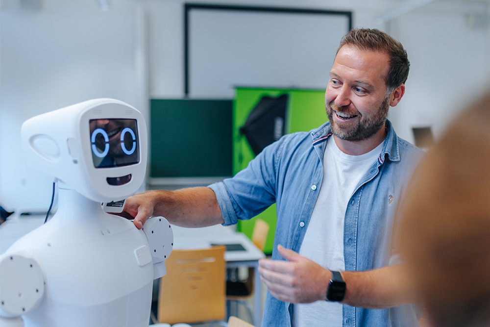 Eine männlich gelesene Person steht neben einem Roboter, dem er eine Hand auf die Schulter gelegt hat. Die Person trägt ein Jeanshemd und ein weißes T-Shirt. Im Hintergrund erkennt man einen Green Screen und professionelles Lichtequipment.