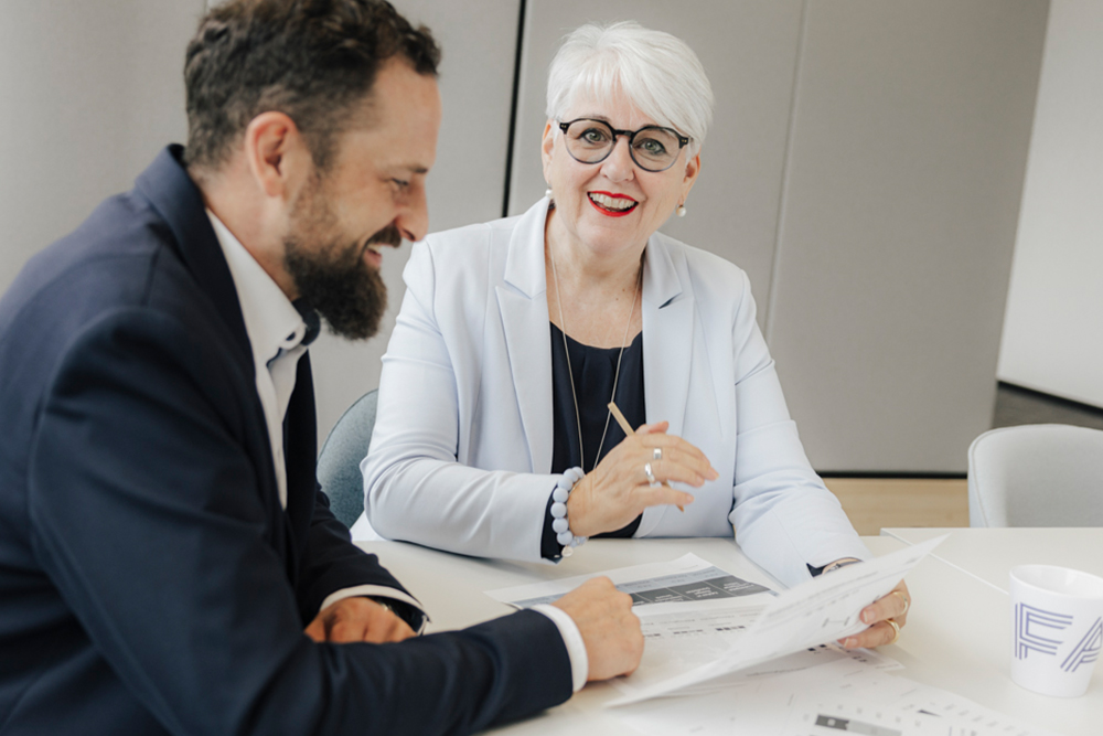Zwei elegant gekleidete Mitarbeitende der FAU Erlangen-Nürnberg sitzen gemeinsam am Tisch und besprechen Dokumente, auf denen sich Zahlen und Statistiken befinden.