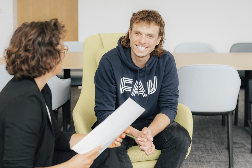 Eine weiblich und eine männlich gelesene Person sitzen in einer modernen Arbeitsumgebung zusammen. Die eine Person lächelt in die Kamera. Sie trägt einen blauen Hoodie mit den drei Buchstaben "FAU".