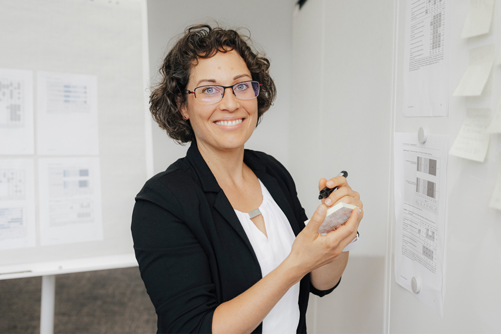 Eine FAU-Mitarbeiterin des Bereichs "Lehre und Studium" präsentiert Evaluationen an einem Whiteboard und lächelt in die Kamera. Sie trägt elegante Kleidung in Schwarz und Weiß.