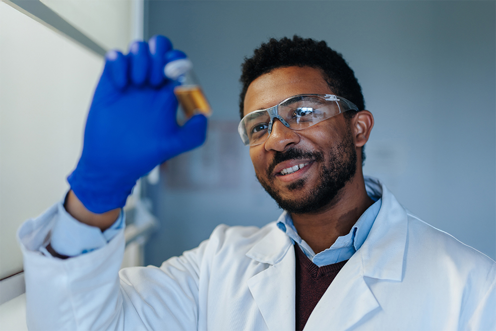 Ein männlich gelesene Person in einem weißen Kittel und mit blauen Laborhandschuhen begutachtet eine wissenschaftliche Probe.