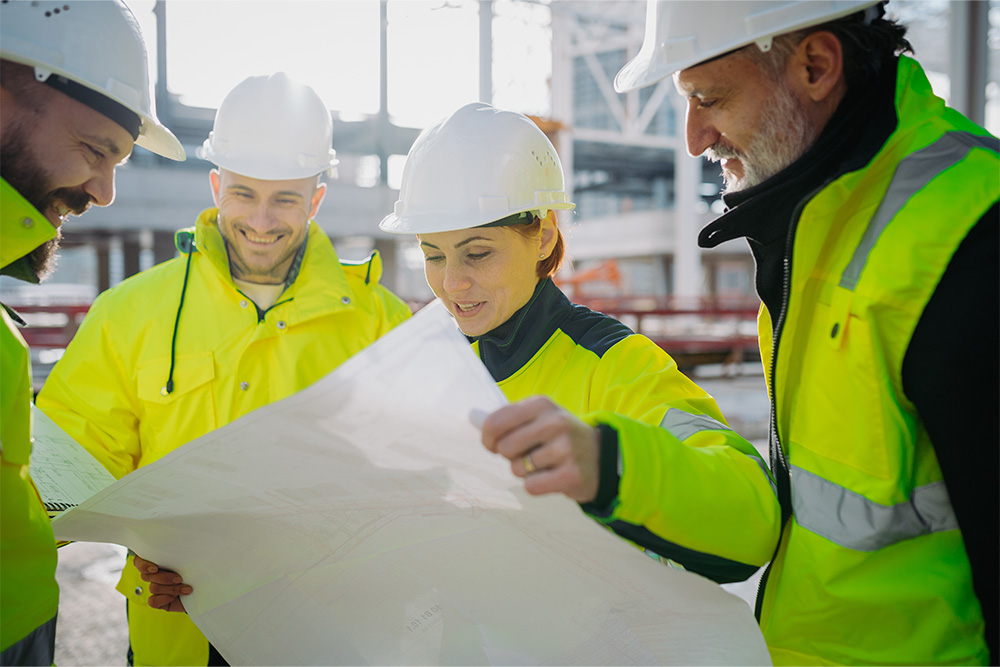Eine Gruppe von Personen steht über einen Bauplan gebeugt zusammen. Alle tragen gelbe Warnwesten sowie weiße Helme. Im Hintergrund sieht man Industriebauten. Alle lächeln freundlich und sind konzentriert auf die Karte.