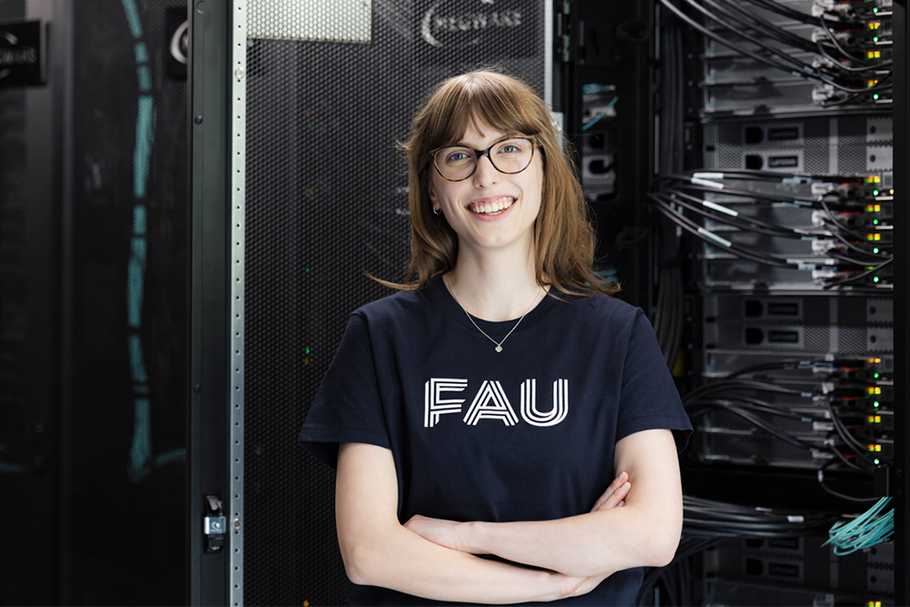 Weiblich gelesene Person steht lächelnd vor einem geöffneten Server-Schrank in einem Serverraum der Friedrich-Alexander-Universität Erlangen-Nürnberg.