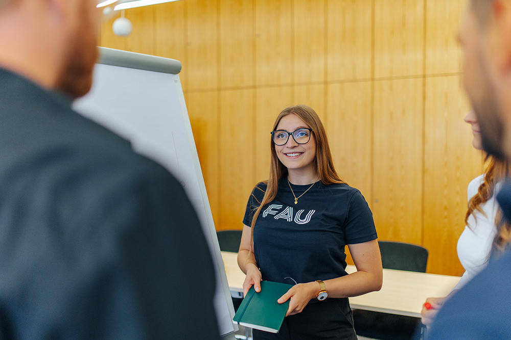 Eine weiblich gelesene Person steht inmitten einer Gruppe von Personen vor einem Flipchart. Sie trägt ein T-Shirt mit dem Logo der Friedrich-Alexander-Universität Erlangen-Nürnberg und hält ein Notizbuch in der Hand. Sie trägt eine Brille und Schmuck. Der Raum im Hintergrund ist vertäfelt.