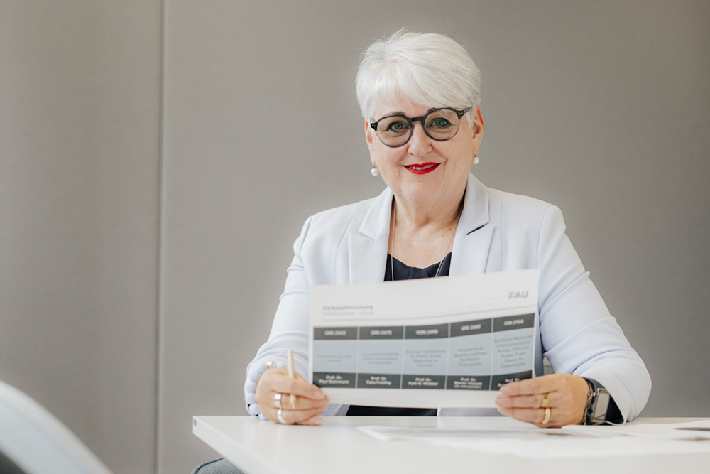 Eine weiblich gelesene Person mit Brille und in eleganter Kleidung sitzt an einem Tisch und liest einen Bericht. Sie trägt Schmuck und lächelt in die Kamera.