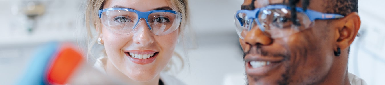 Zwei Auszubildende als Chemielaborant/-innen stehen nebeneinander in einem Labor. Die eine Person hält eine Probe in die Kamera. Beide Personen tragen einen weißen Kittel und Schutzbrillen.