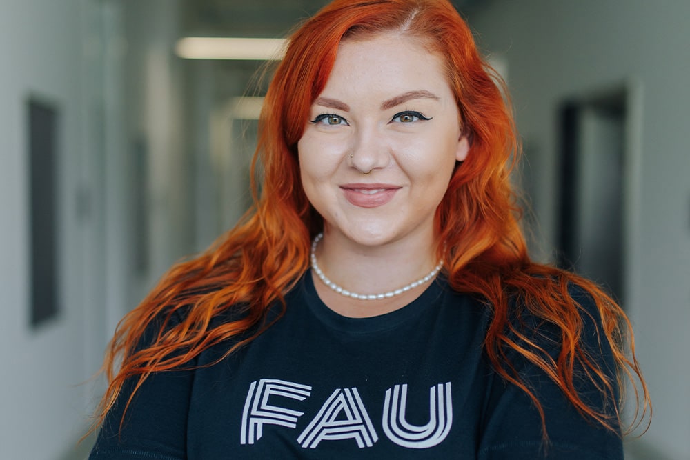 Eine weiblich gelesene Person mit roten welligen Haaren lächelt in die Kamera. Sie trägt ein T-Shirt mit dem Logo der Friedrich-Alexander-Universität Erlangen-Nürnberg, eine Perlenkette und Nasenpiercings.