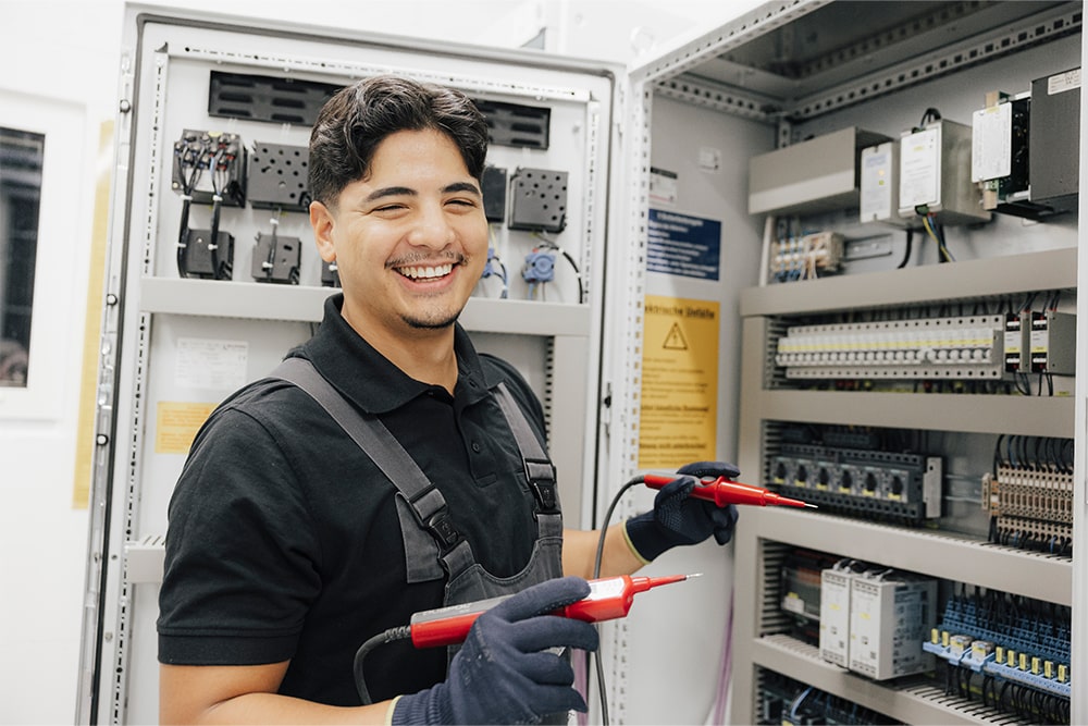 Auszubildender für den Bereich Elektronik steht an einem Elektroschrank, an dem er eine Messung vornehmen möchte. Er trägt eine Latzhose, Handschuhe und lächelt in die Kamera.
