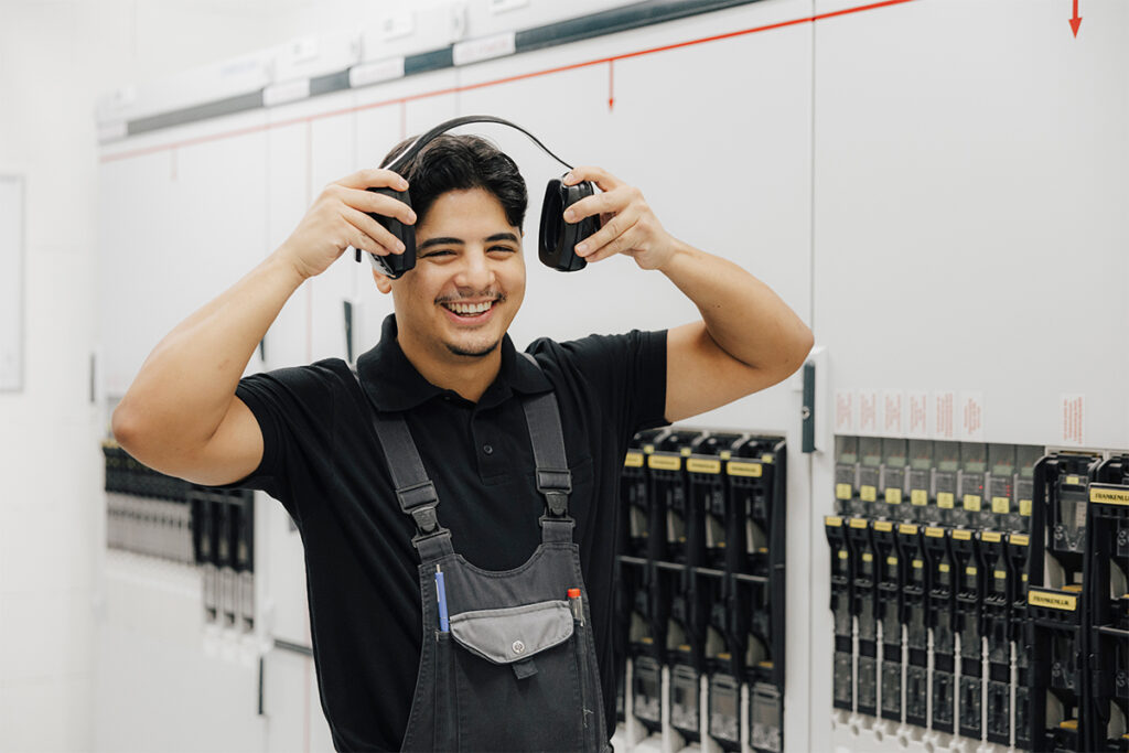 Männlich gelesene Person in einem Elektroraum mit Elektroschrank trägt eine Latzhose und ist gerade im Begriff, sich einen Hörschutz anzuziehen. Dabei lächelt die Person in die Kamera.