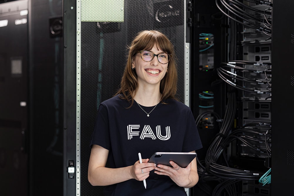 Eine Auszubildende als Fachinformatikerin für Systemintegration trägt ein blaues T-Shirt mit dem Logo der Friedrich-Alexander-Universität Erlangen-Nürnberg. Sie steht mit einem Tablet in der Hand in einem Serverraum.