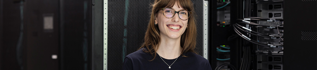 Eine Auszubildende als Fachinformatikerin für Systemintegration trägt ein blaues T-Shirt mit dem Logo der Friedrich-Alexander-Universität Erlangen-Nürnberg. Sie steht mit einem Tablet in der Hand in einem Serverraum.