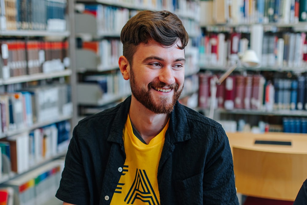Männlich gelesene Person sitzt in einer Bibliothek. Die Person trägt ein blaues Hemd über einem gelben T-Shirt und lächelt eine Person außerhalb der Kamer an.