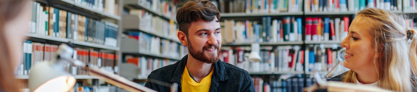 Zwei Personen sitzen in einer Bibliothek. Die linke Person trägt ein blaues Hemd über einem gelben T-Shirt und lächelt die andere Person an.