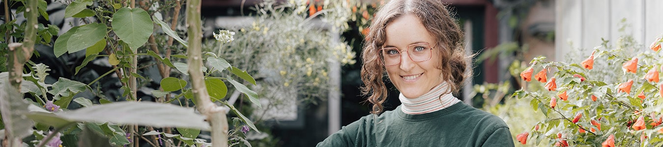Auszubildende steht in einem Gewächshaus vor verschiedenen Pflanzentöpfen, Sie hält eine Pflanze in der Hand, die sie gerade beschneidet.