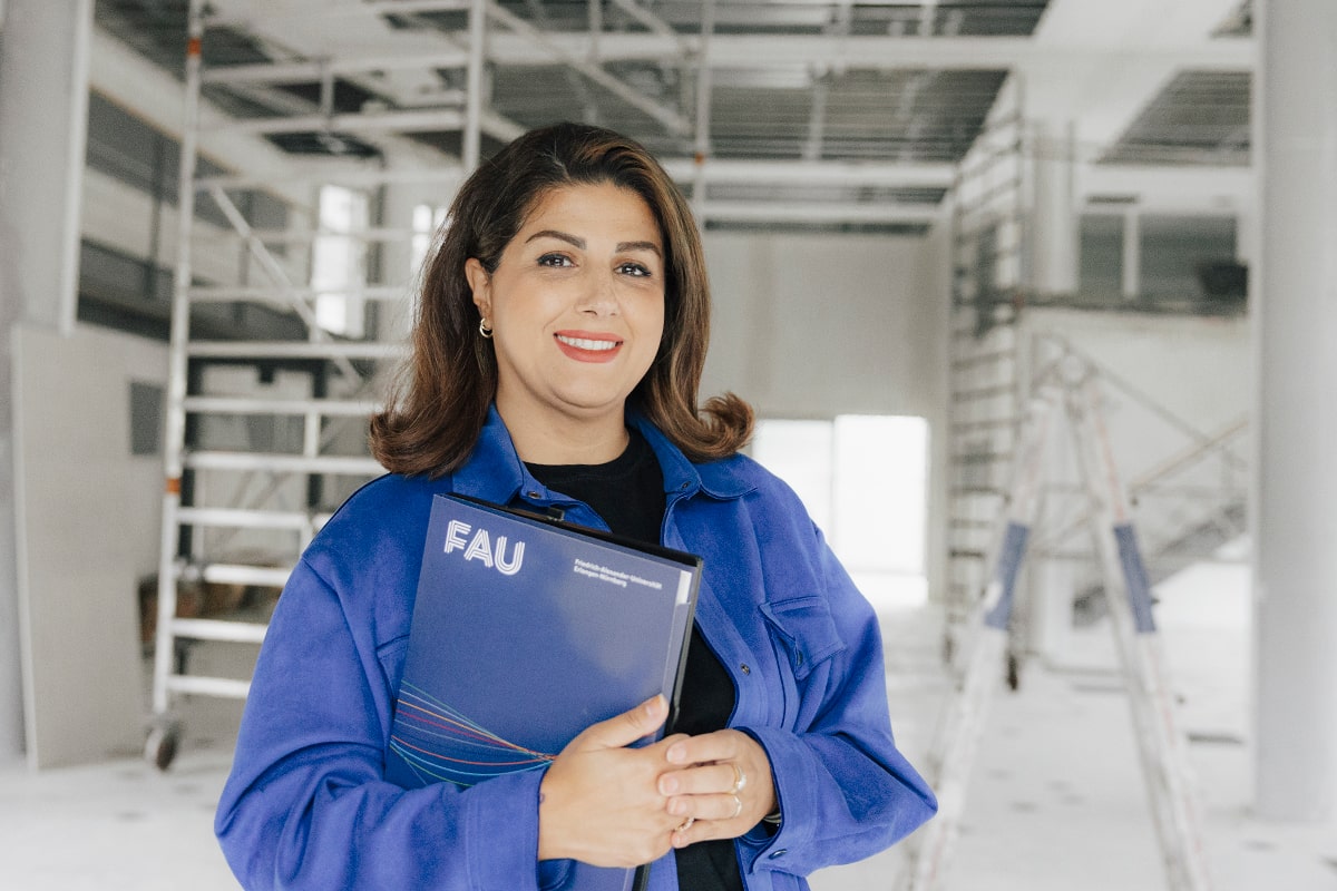 Weiblich gelesene Person steht auf einer Indoor-Baustelle. Sie trägt eine Mappe mit dem Logo der Friedrich-Alexander-Universität Erlangen-Nürnberg und lächelt in die Kamera. Im Hintergrund sieht