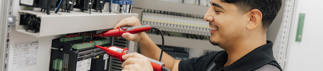 Eine männlich gelesene Person steht an einem Elektroschrank und nimmt dort eine Messung vor.