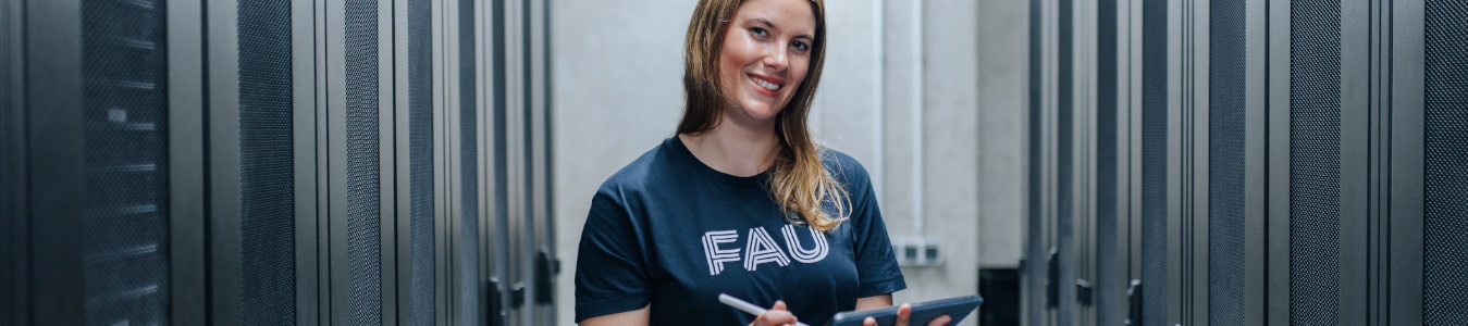 Eine weiblich gelesene Person steht in einem Serverraum. Sie hält ein Tablet in der Hand und lächelt in die Kamera.