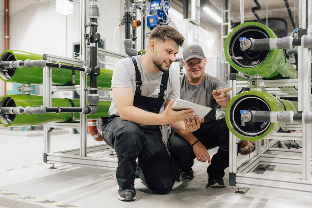 Zwei männlich gelesene Personen knien zwischen zwei Anlagensystemen in einem modernen Gebäudeteil der Friedrich-Alexander-Universität Erlangen-Nürnberg. Die eine Person trägt eine Latzhose und hält ein Tablet in der Hand. Die andere Person trägt eine Käppi und deutet auf einen Punkt an einer der Anlagen.