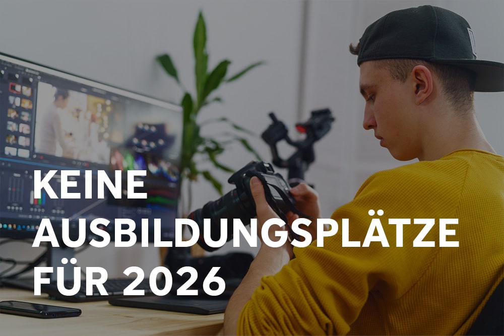 Auszubildender sitzt vor einem PC, auf dem ein Schnittprogramm geöffnet ist und betrachtet währenddessen Bilder auf einer Kamera, die er in der Hand hält. Auf dem Bild steht "Keine Ausbildungsplätze für 2026".