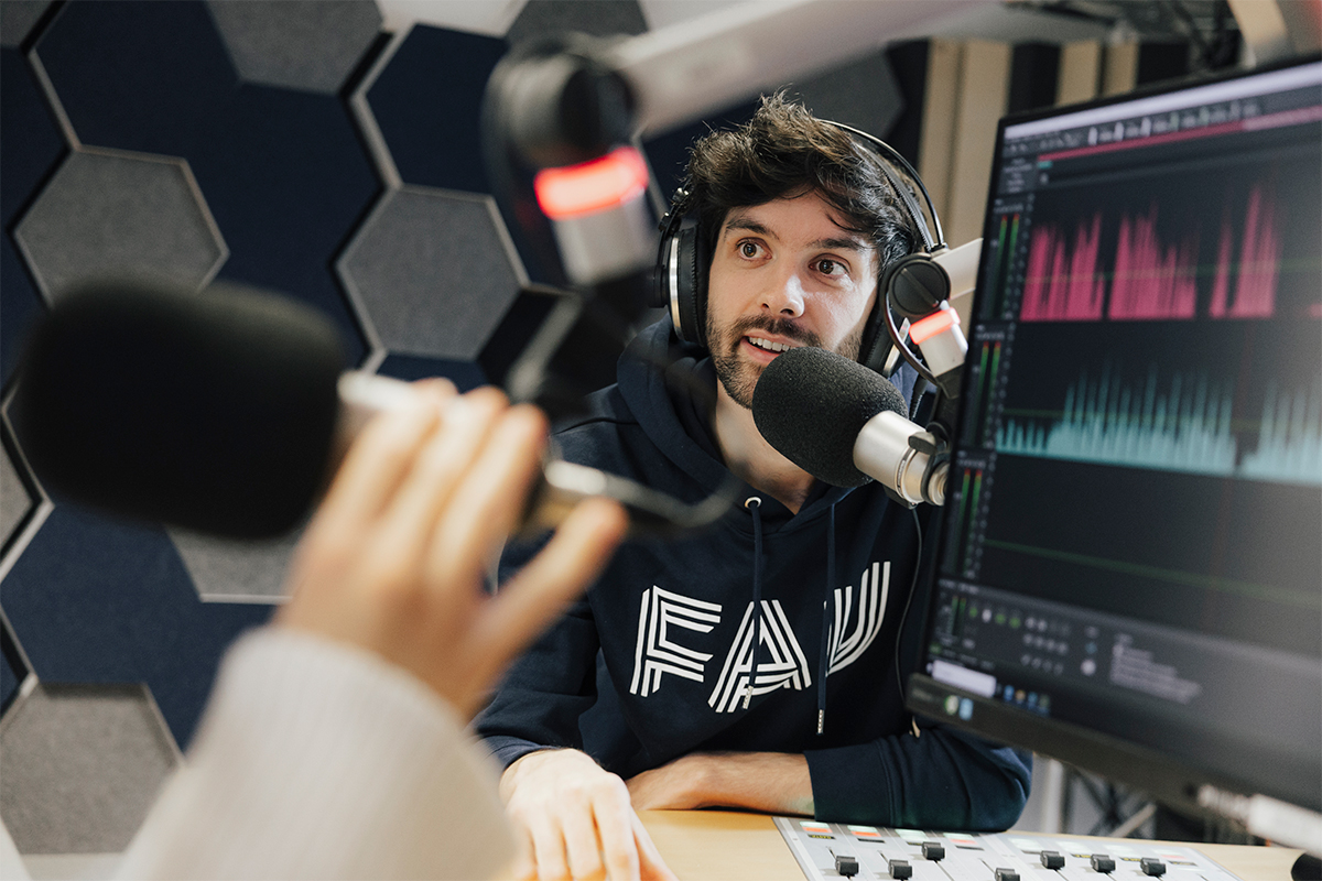 Männlich gelesene Person steht in einem Tonstudio an einem Mikrofon. Die Person trägt einen Pullover mit dem Logo der Friedrich-Alexander-Universität Erlangen-Nürnberg (FAU) sowie Kopfhörer. Im Hintergrund sieht man die Kacheln einer Schallisolationswand. Im Vordergrund steht ein Bildschirm mit einer bunten Tonanzeige.