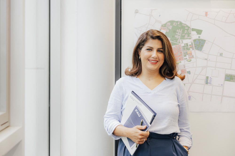Eine weiblich gelesene Person steht vor einem Gebäudeplan. Sie trägt Business-Kleidung und hält einen FAU-Block in der Hand.