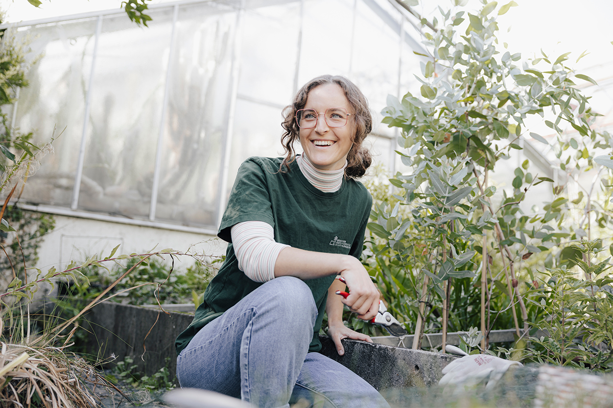 Weiblich gelesene Person kniet vor einem Gewächshaus neben einem Beet. Die Person trägt eine Brille und ein grünes T-Shirt mit dem Logo des Botanischen Gartens Erlangen und hält eine Gartenschere in der Hand. Sie lächelt.