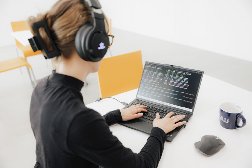Weiblich gelesene Person sitzt an einem Laptop, auf dem ein Code abgebildet ist. Auf dem Tisch steht zudem eine Tasse mit dem Logo der Friedrich-Alexander-Universität Erlangen-Nürnberg. Die Person trägt eine Brille, Kopfhörer und ein schwarzes Langarmshirt.