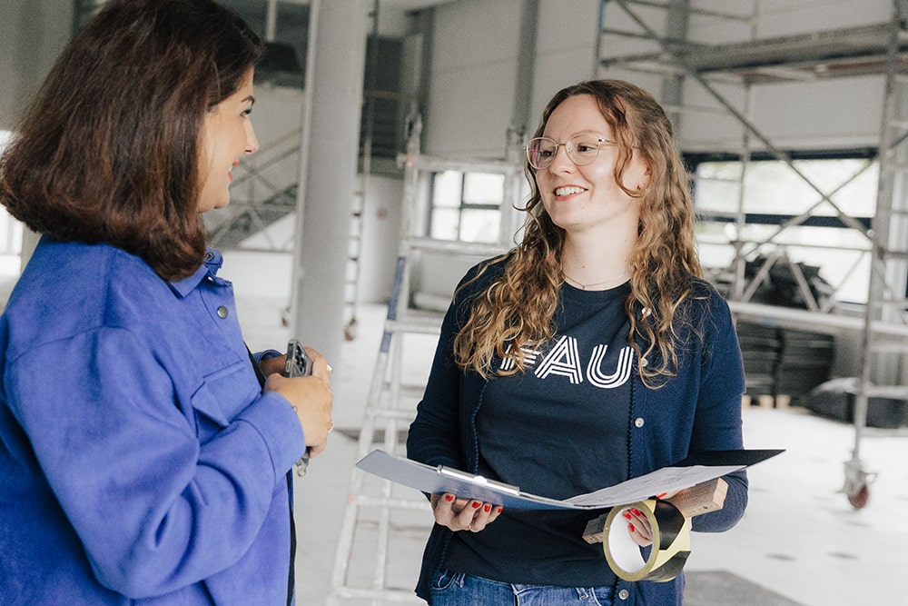Zwei weiblich gelesene Personen stehen auf einer Baustelle zusammen. Die eine Person hält einen Ordner mit Unterlagen und einen Maßstab in den Händen, die andere Person trägt ein blaues Hemd und hält ein Smartphone in der Hand.